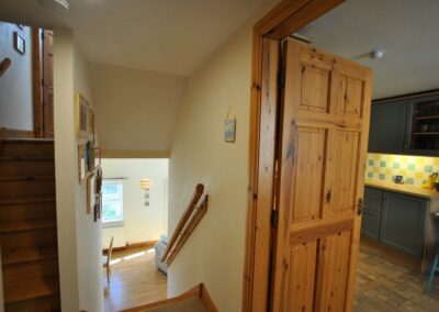 View from top of wooden staircase. On the right is an open door into the kitchen. On the left is another staircase leading upwards.