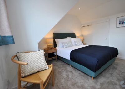 Double bed between bedside tables and lamps with an inbuilt storage cupboard beyond. In the foreground a chair sits beneath a window.