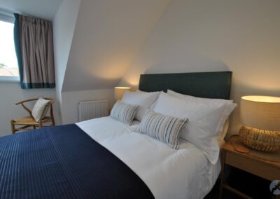 Double bed with navy and white bedding. There is a window with a chair beneath to the left.