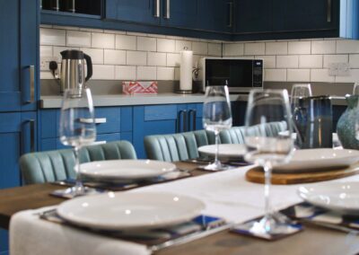 Dining table set with crockery and wine glasses, looking over to a microwave in the corner of the kitchen beneath blue cupboards.